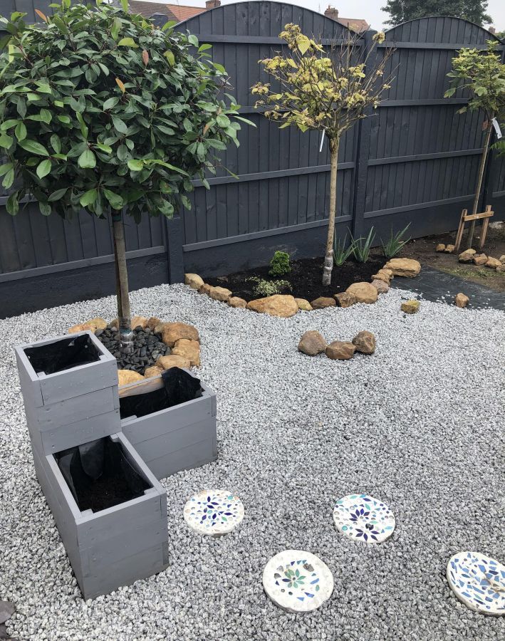 Modern garden with grey gravel, black fence, young trees, and mosaic stepping stones.