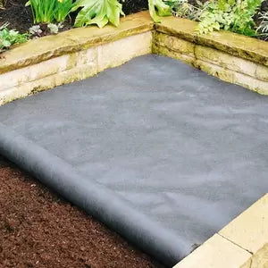 A garden bed covered with black landscape fabric to prevent weed growth, surrounded by stone borders.